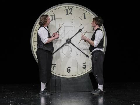 Standbild aus dem Theaterstück "Eine Minute". Zwei männliche Schauspieler stehen vor einer großen Uhr und schauen sich an.